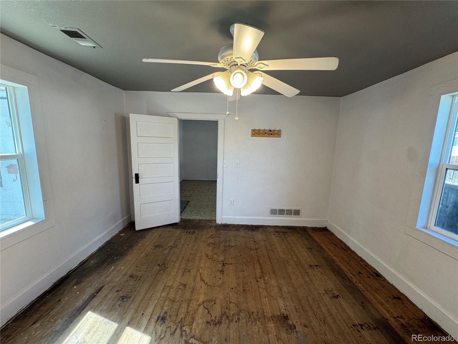Empty room with worn wood flooring, a ceiling fan, and windows on two walls.