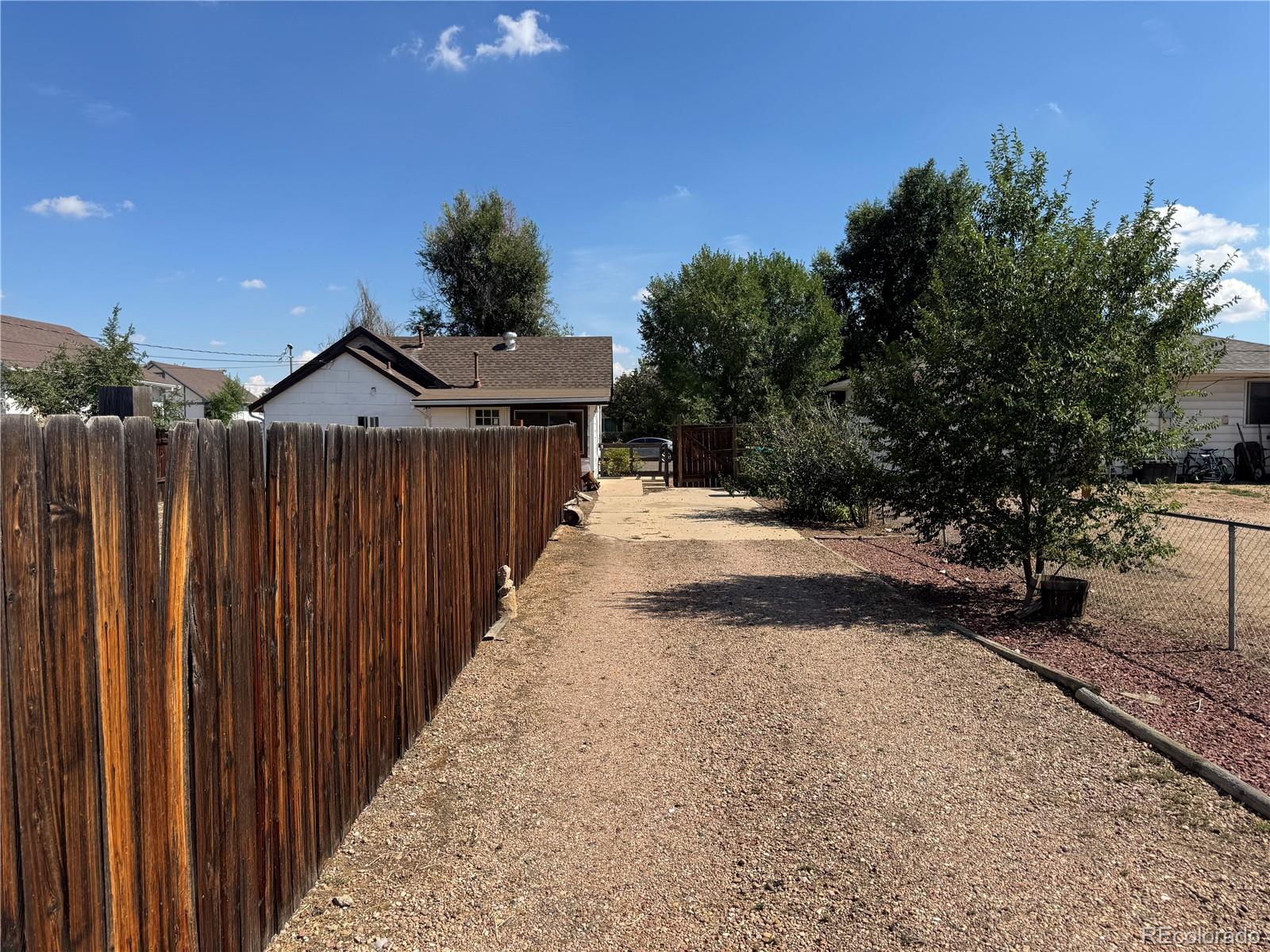 Gravel backyard path leading to the rear of a house, bordered by a tall wooden fence and trees.