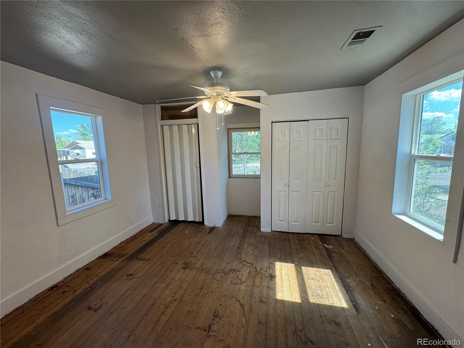 Empty room with worn wood floors, three windows, a ceiling fan, and a sliding-door closet.