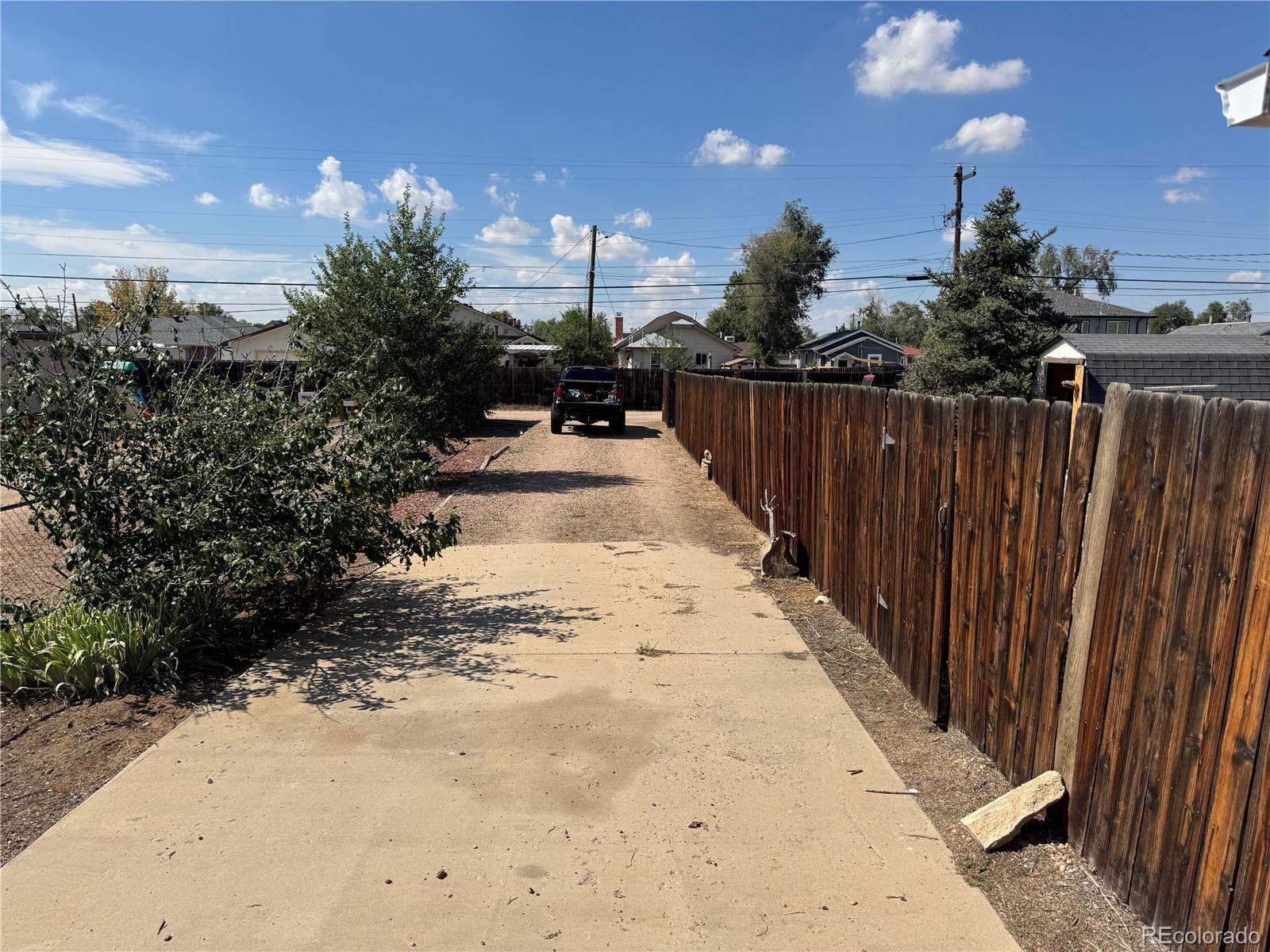 Concrete driveway leading to a gravel yard, bordered by a tall wooden fence and shrubs.