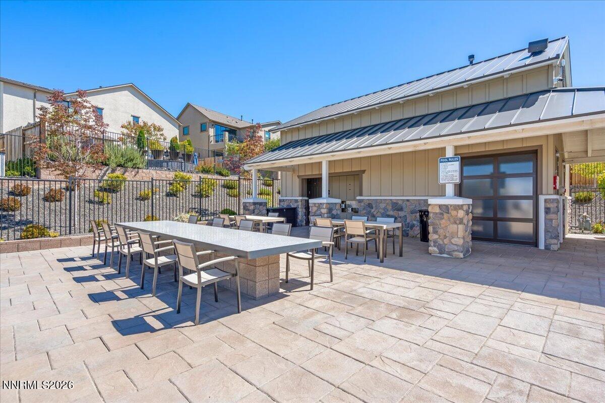 Outdoor poolside patio with dining tables beside a stone-clad facility and landscaped homes.