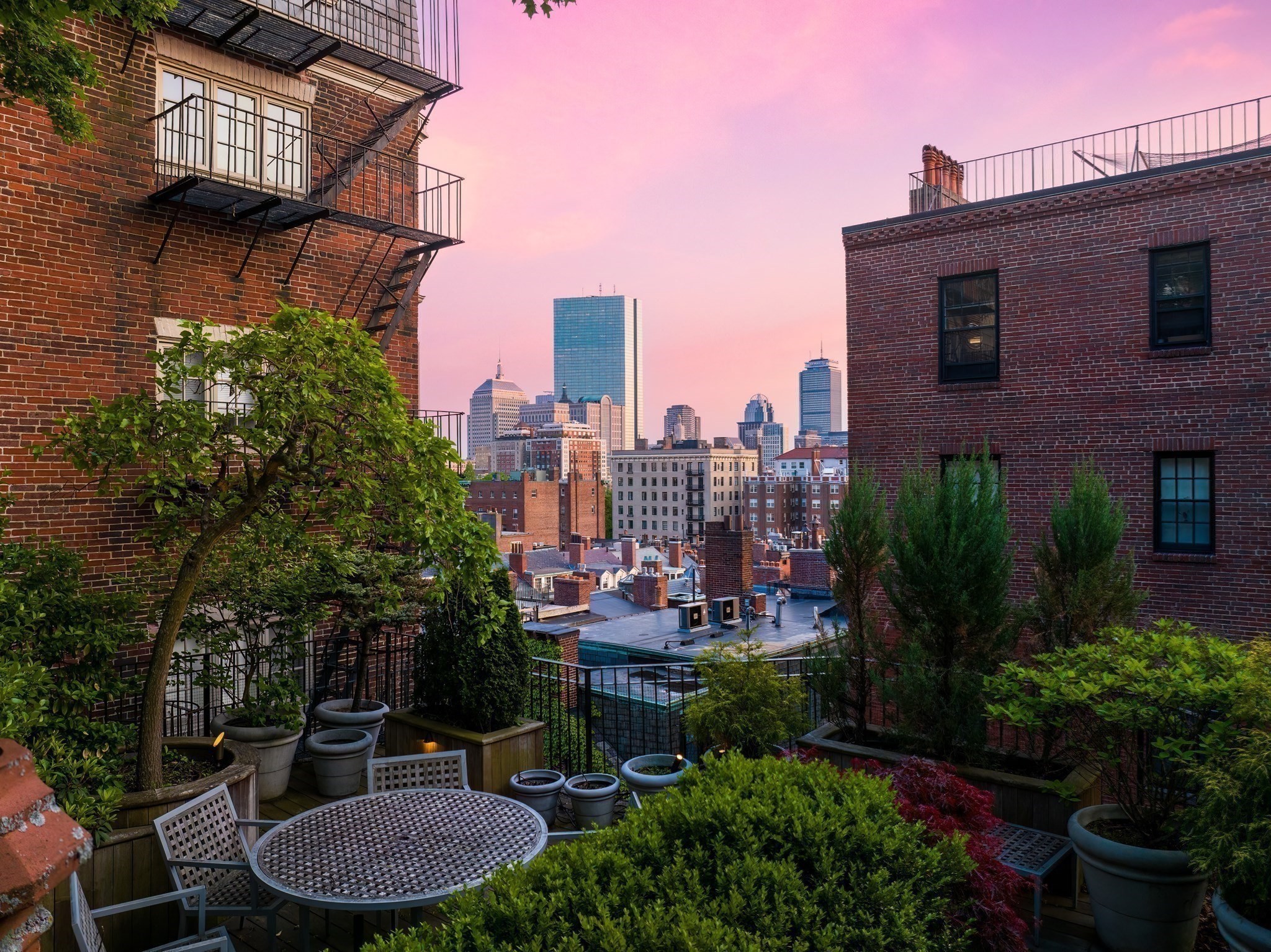 Rooftop terrace with potted greenery and dining set overlooking the Boston skyline at dusk.