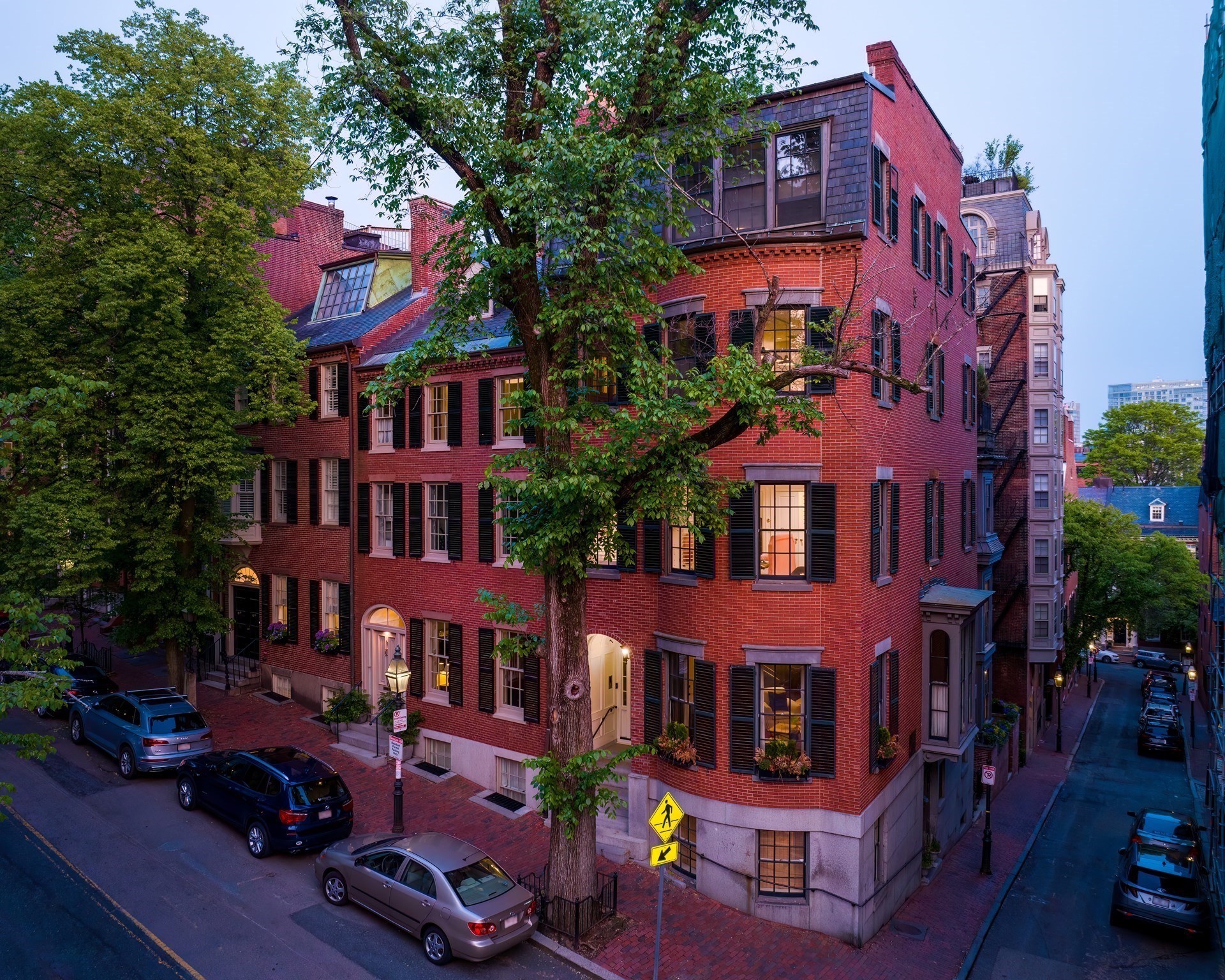 Red-brick corner townhouse with black shutters and a large tree in front at dusk.