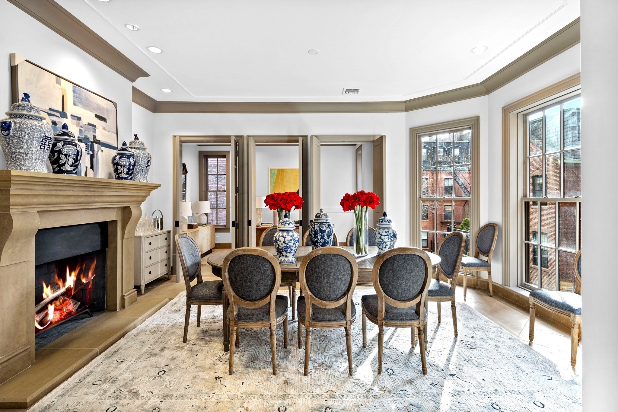 Dining room with a lit stone fireplace, tall windows, and a table set with porcelain vases and red flowers.