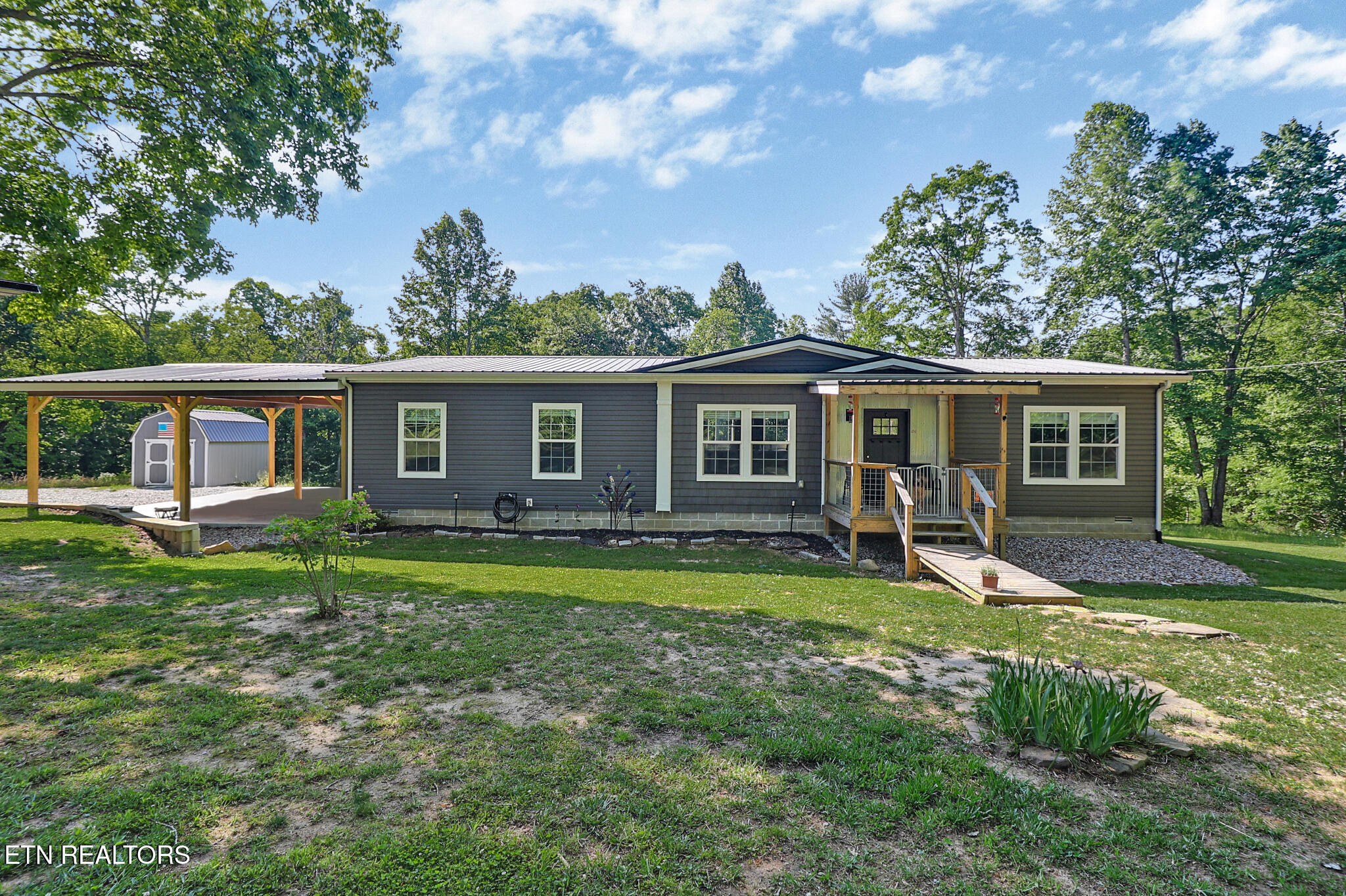 Gray single-story home with a covered porch, long carport, and small shed in a wooded yard.