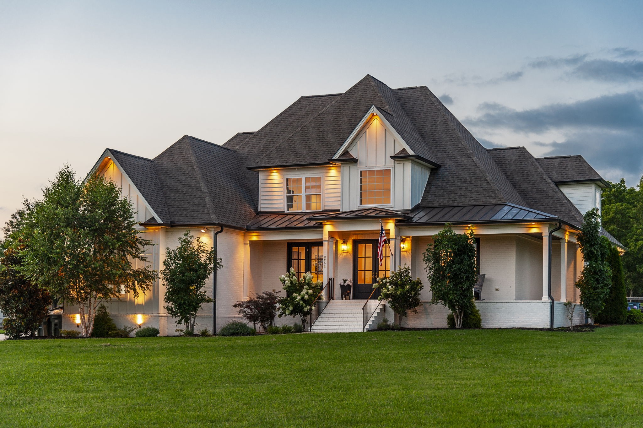 White two-story home with steep gabled roof, illuminated front porch, and broad green lawn at dusk.