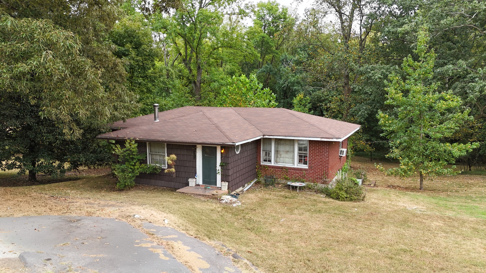 Single-story brick and shingle house surrounded by mature trees, with a wide roof and dry lawn.