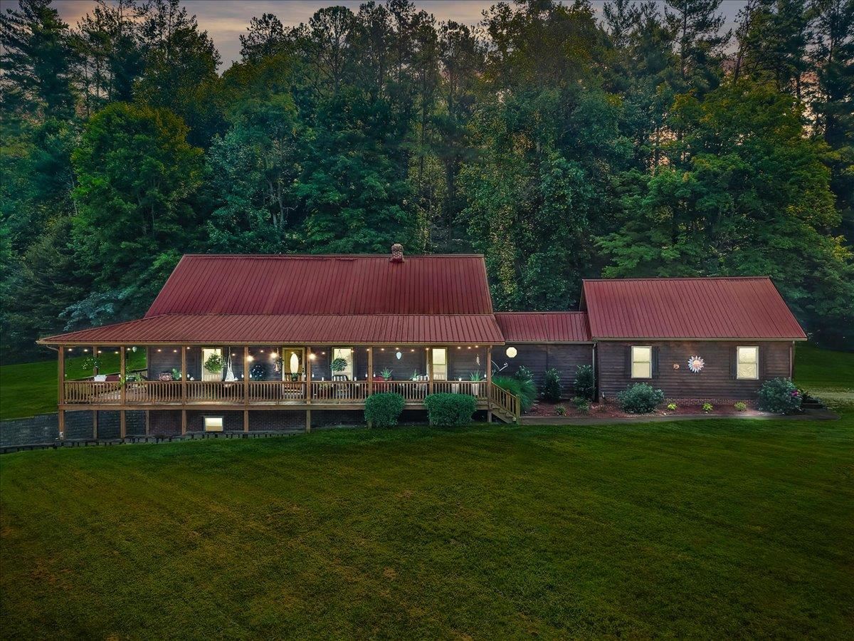 Wood-sided home with a red metal roof, broad covered porch, and wooded backdrop.