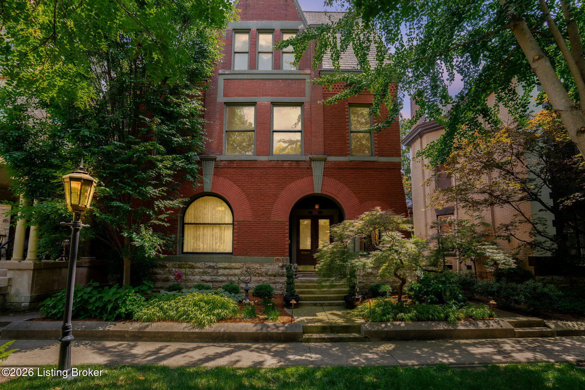 Red brick historic home with arched entry, mature trees, and landscaped front garden.