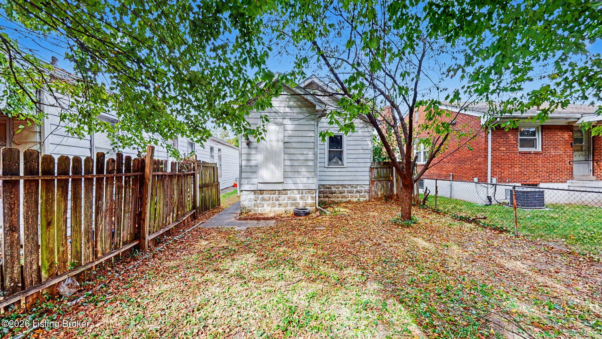 Rear of a gray house and leaf-covered backyard beneath mature trees.
