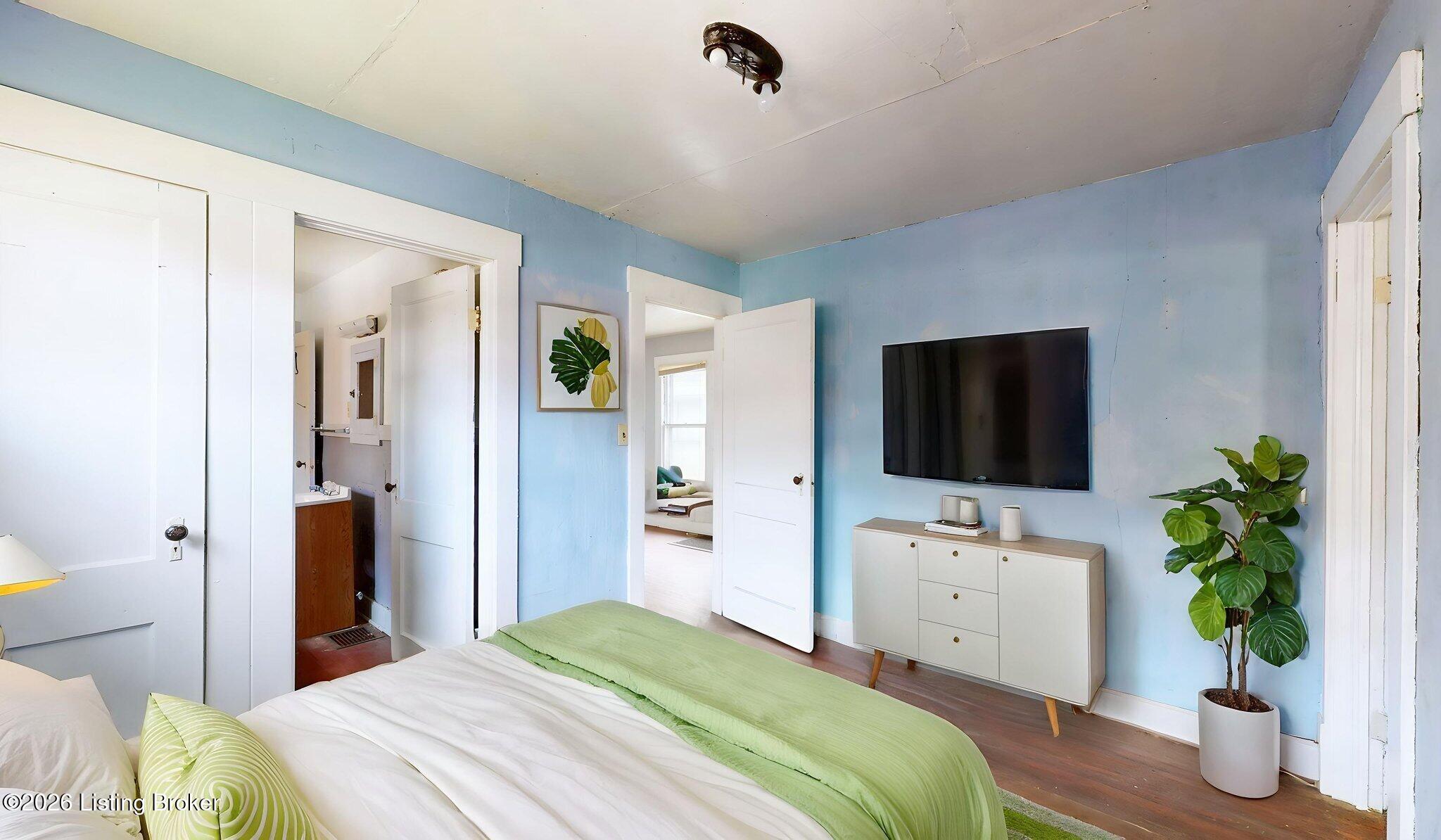Bedroom with light blue walls, green-and-white bedding, and a wall-mounted TV above a dresser.