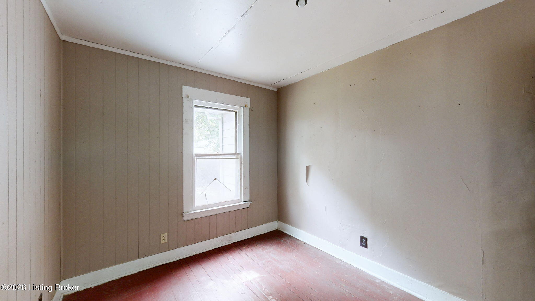 Empty room with reddish plank flooring, paneled walls, and a single window.