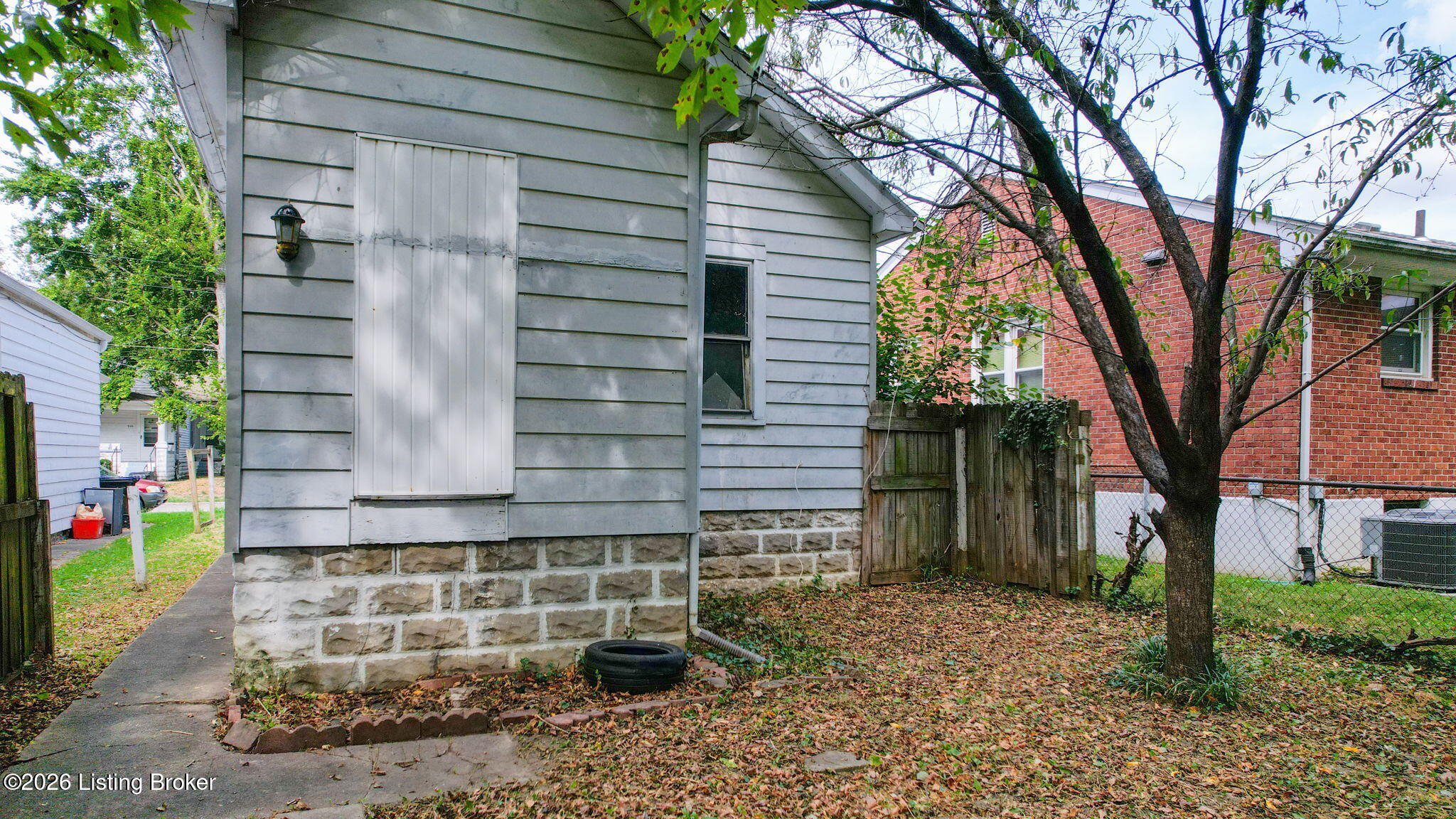 Gray-sided house with a stone foundation beside a leaf-covered yard and mature tree.