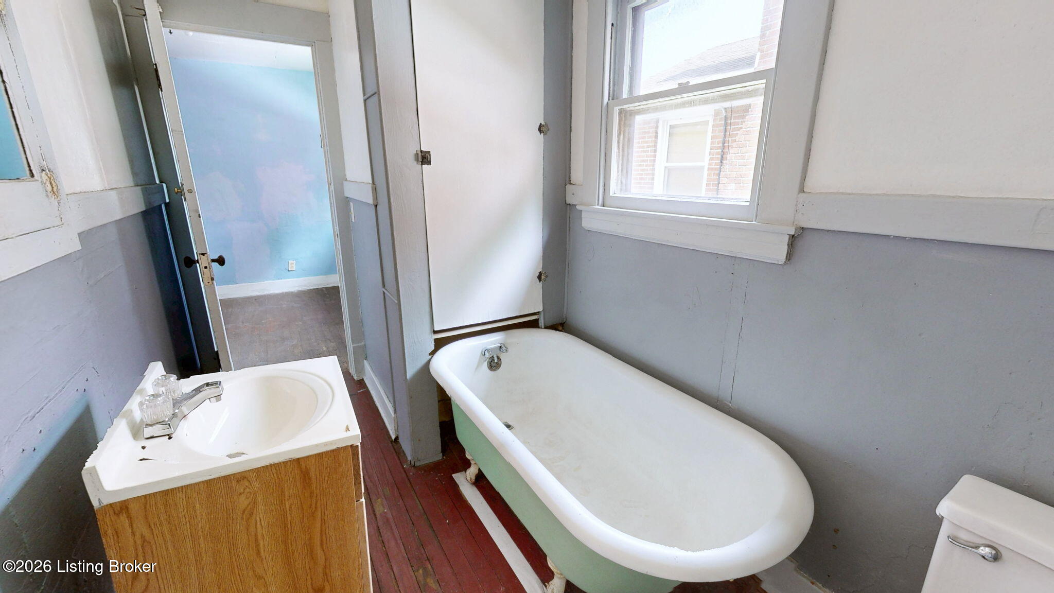 Bathroom with a white-and-green freestanding tub, wood-front sink vanity, and worn red-painted floorboards.