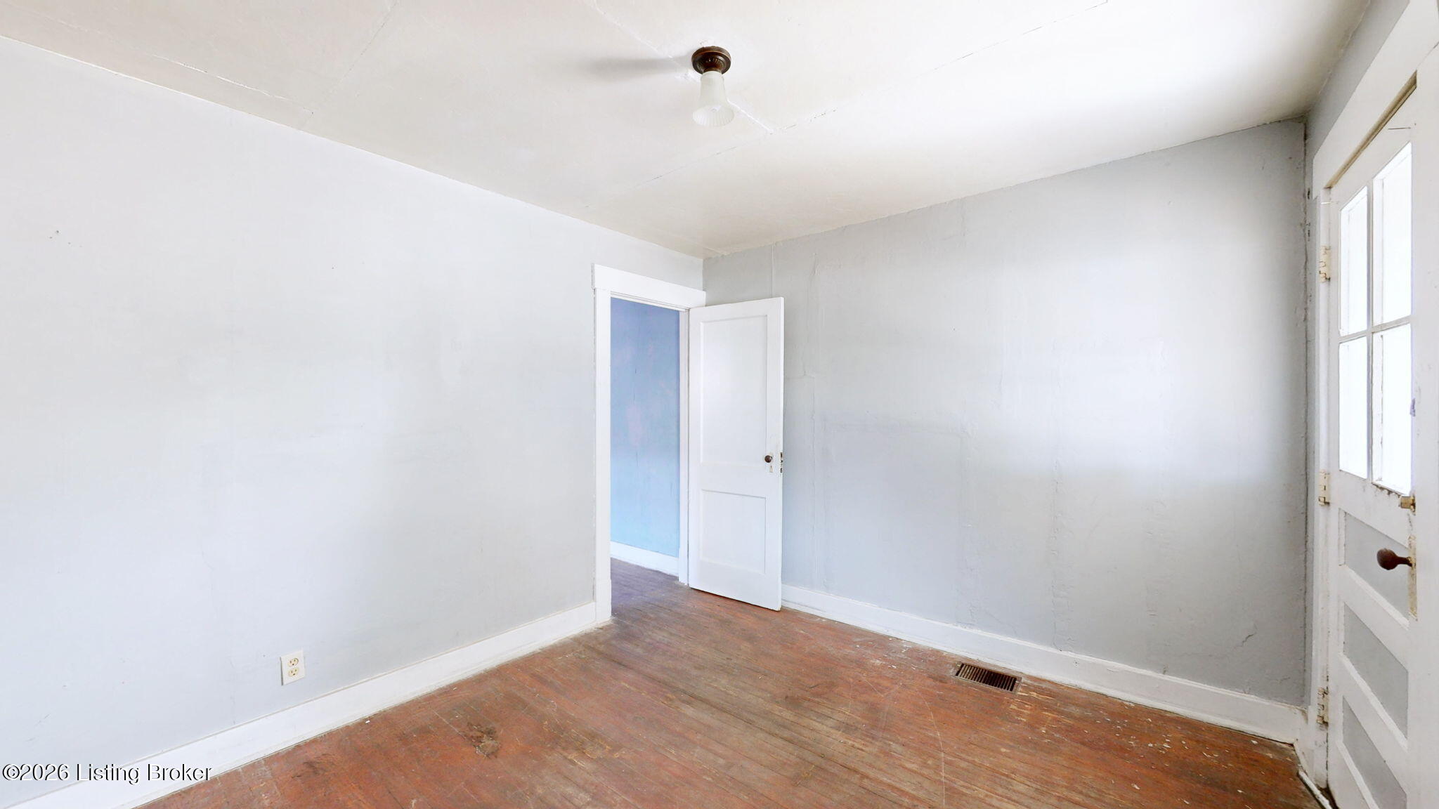 Empty room with worn hardwood floors, pale gray walls, and an open doorway to a blue room.