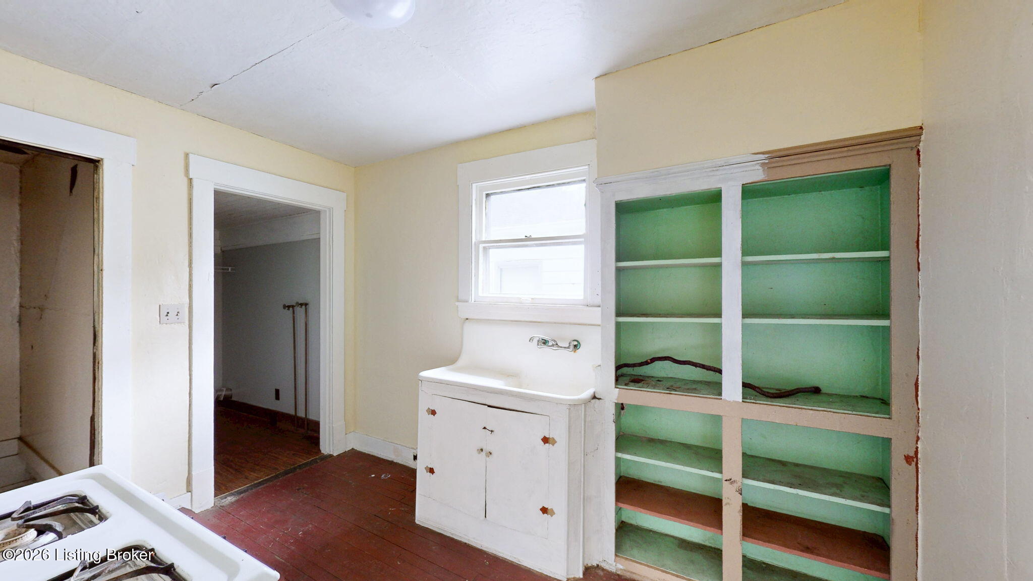 Older kitchen with built-in green shelves, a white sink cabinet, and red-painted wood floor.