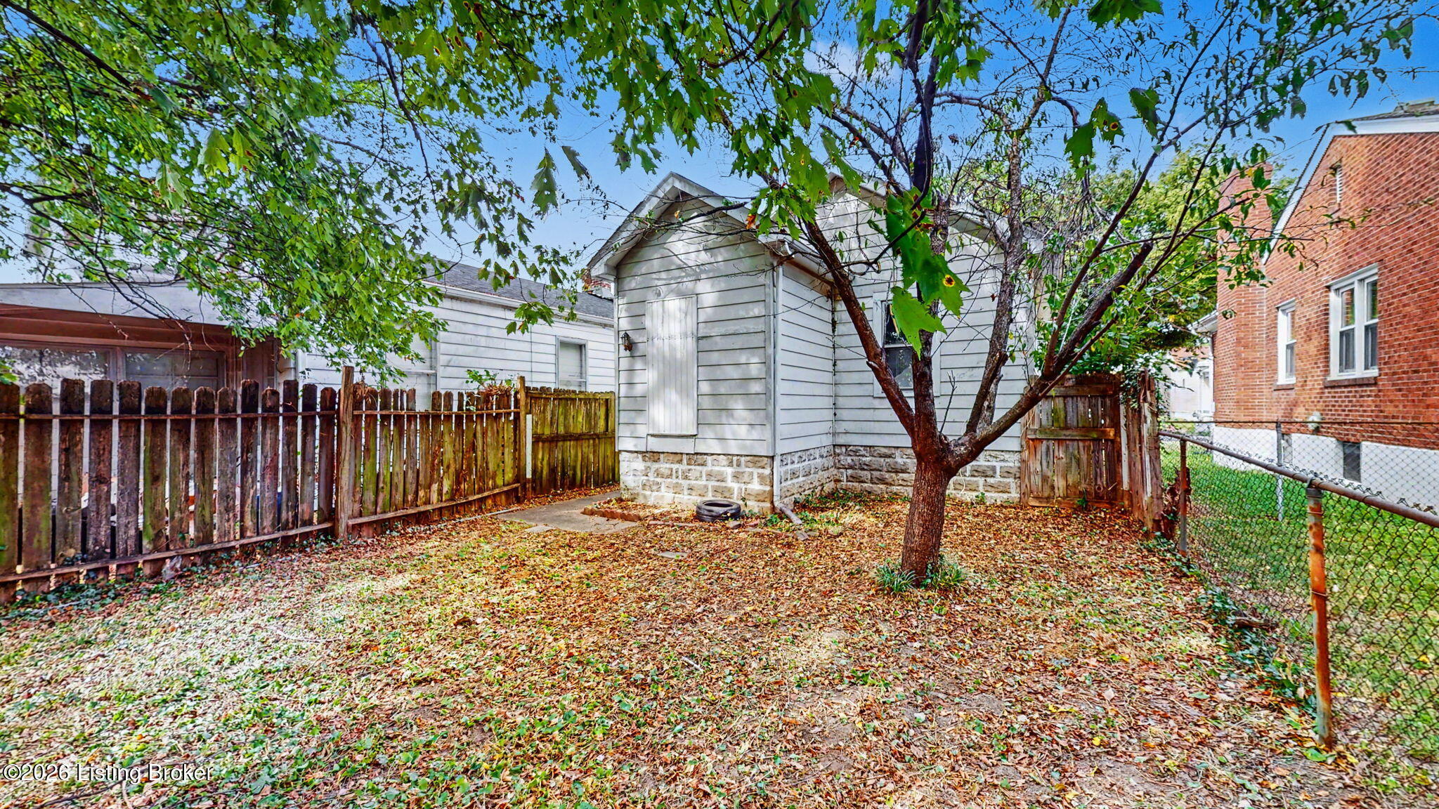 Fenced backyard with a mature tree, fallen leaves, and a small white-sided building.