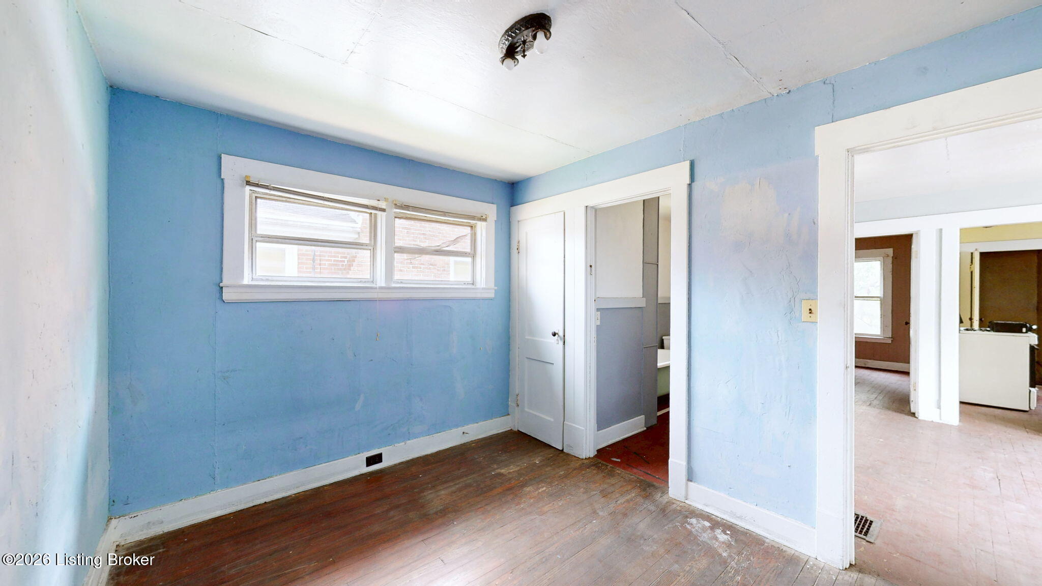 Empty blue room with worn hardwood floors, two windows, and open doorways to adjoining rooms.