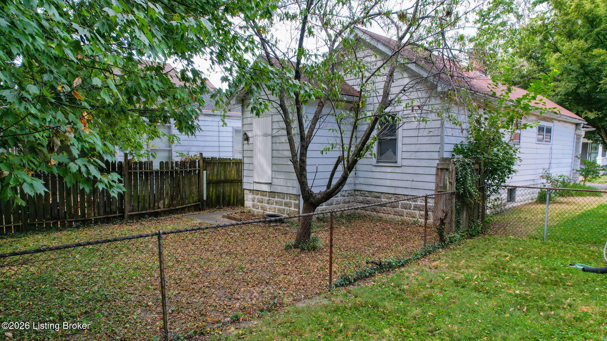 Side of a light-colored house with a red roof, mature trees, and fenced yard.