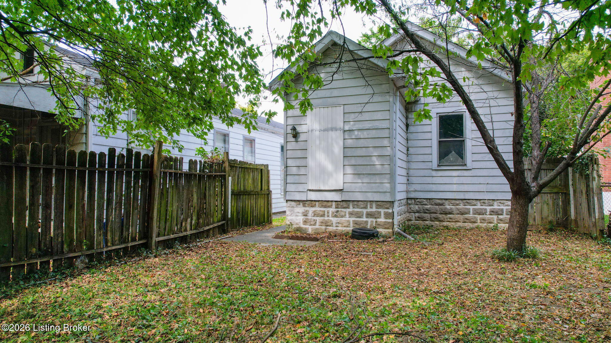House viewed from a fenced backyard, with a stone foundation, mature trees, and fallen leaves.
