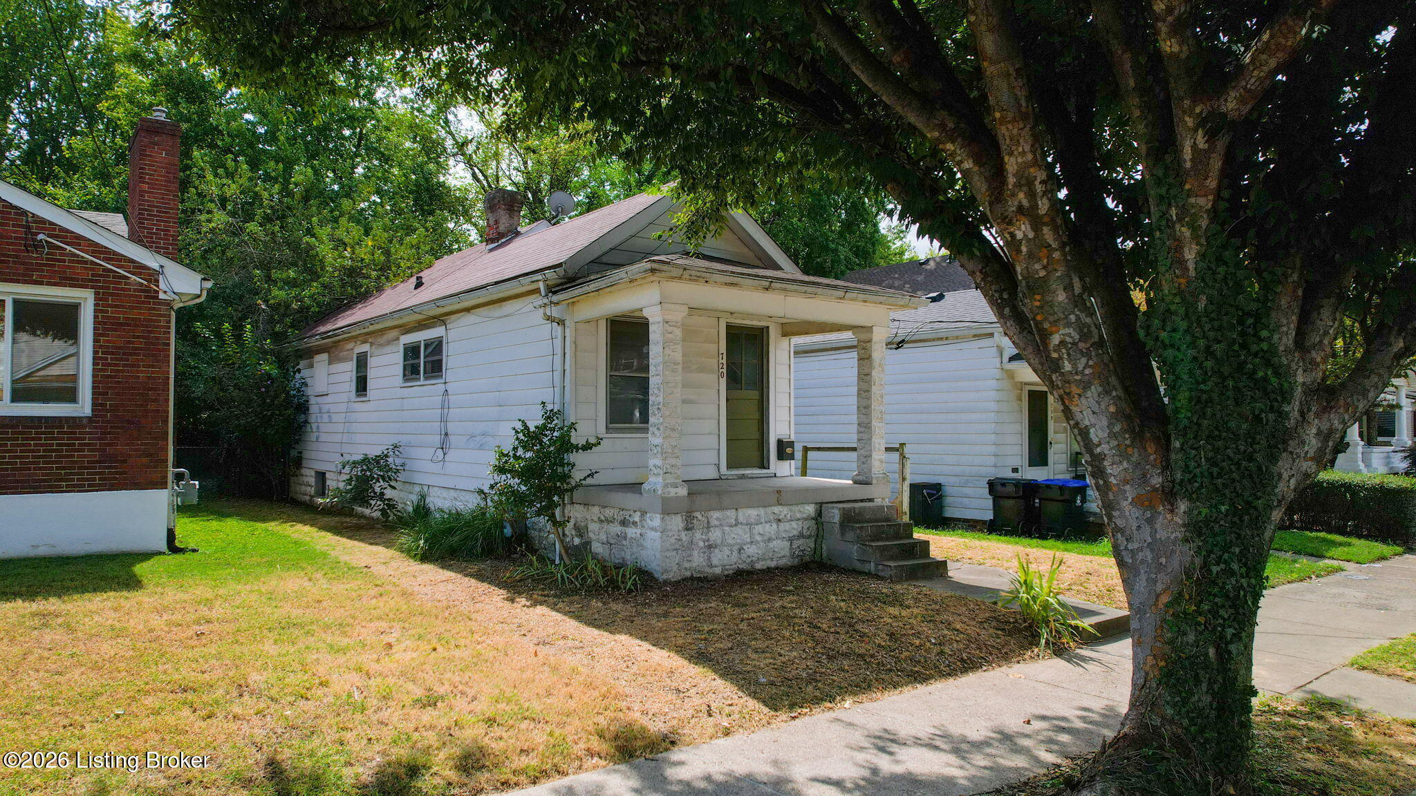 White-sided house with a small raised porch and stone steps, shaded by a large mature tree.