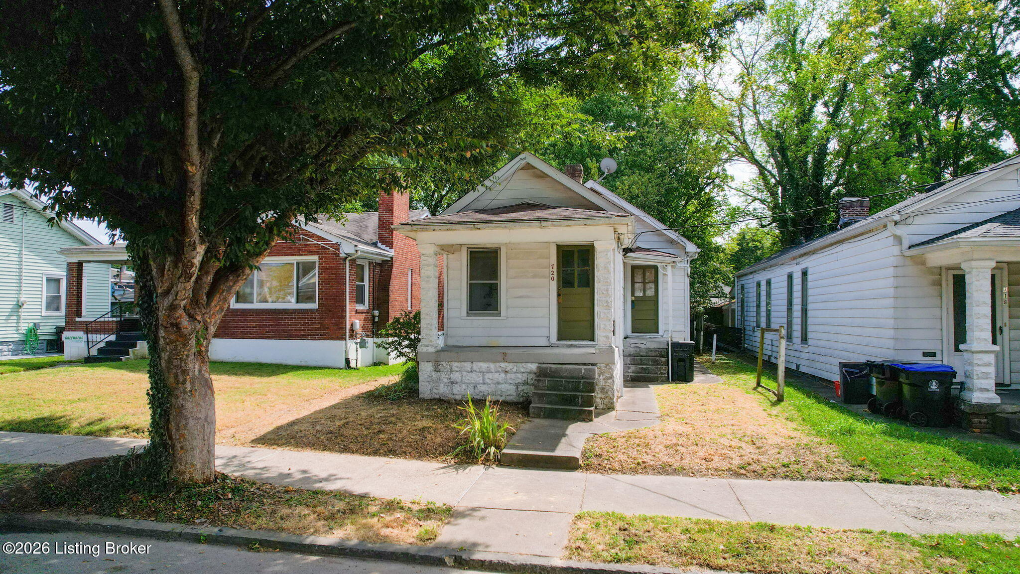Small white house with a stone-front porch and steps, partly shaded by a large tree.