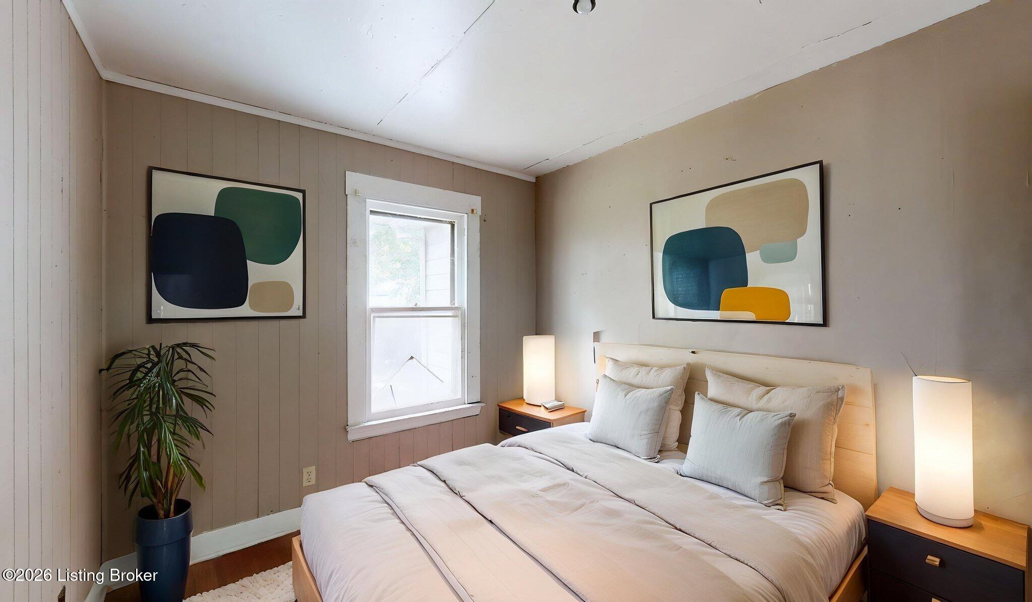 Furnished bedroom with pale bedding, abstract wall art, a window, and a potted plant.