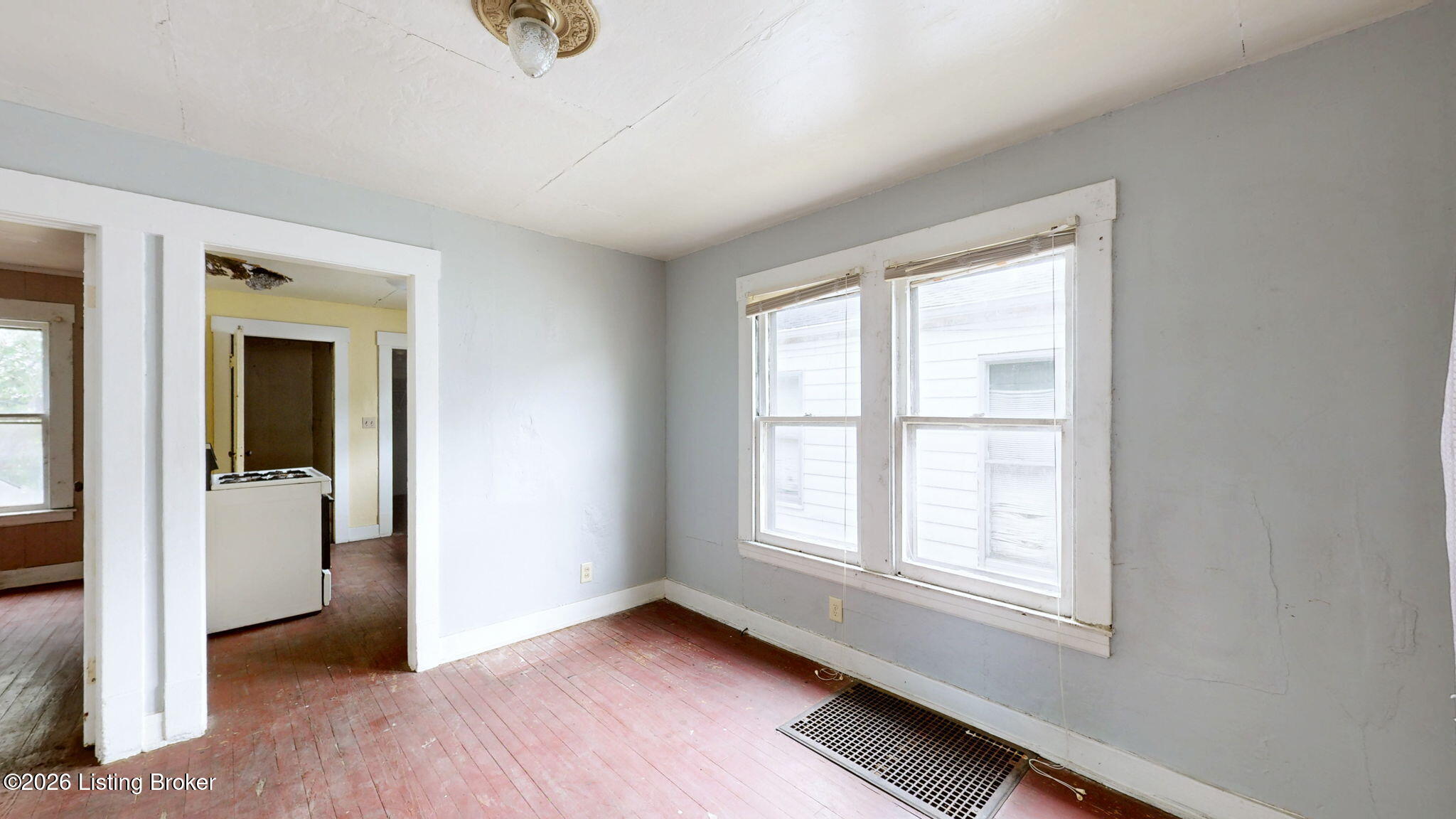 Empty room with red-toned wood floors, two windows, and a large floor register.
