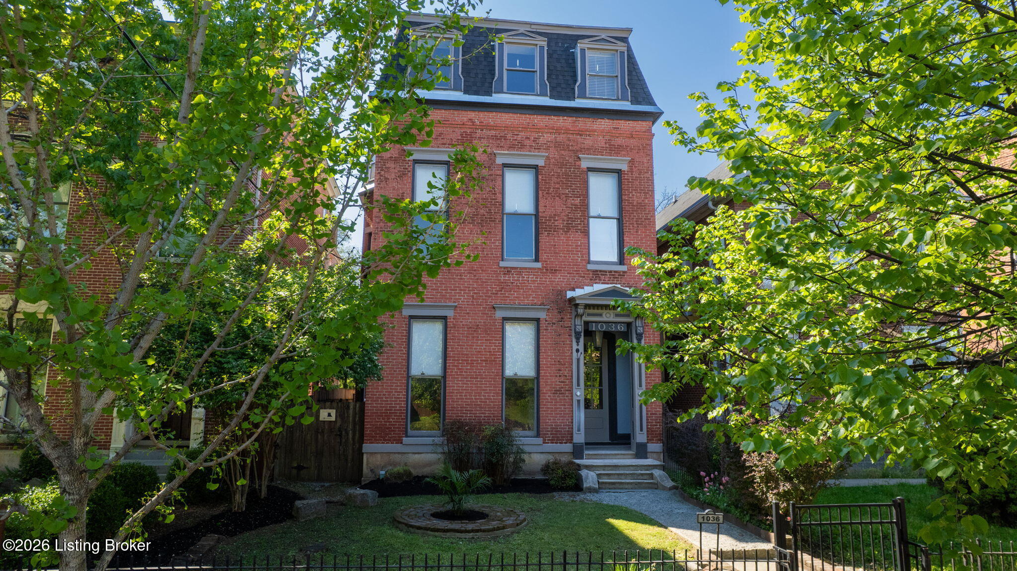 Historic red-brick house with a mansard roof, framed by mature trees and a landscaped front yard.