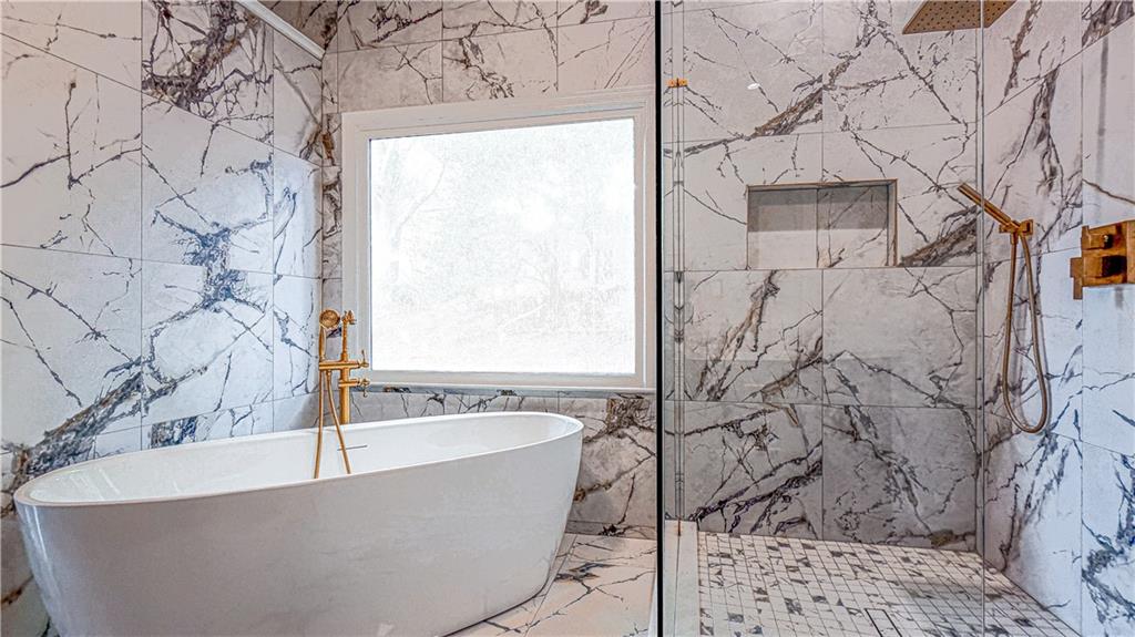 Marble bathroom with a freestanding soaking tub, large window, and glass-enclosed shower with gold fixtures.