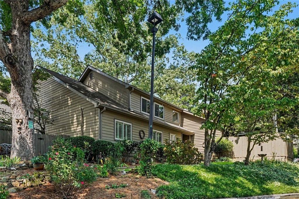 Two-story taupe-sided house behind a leafy landscaped yard with mature trees and a wooden fence.