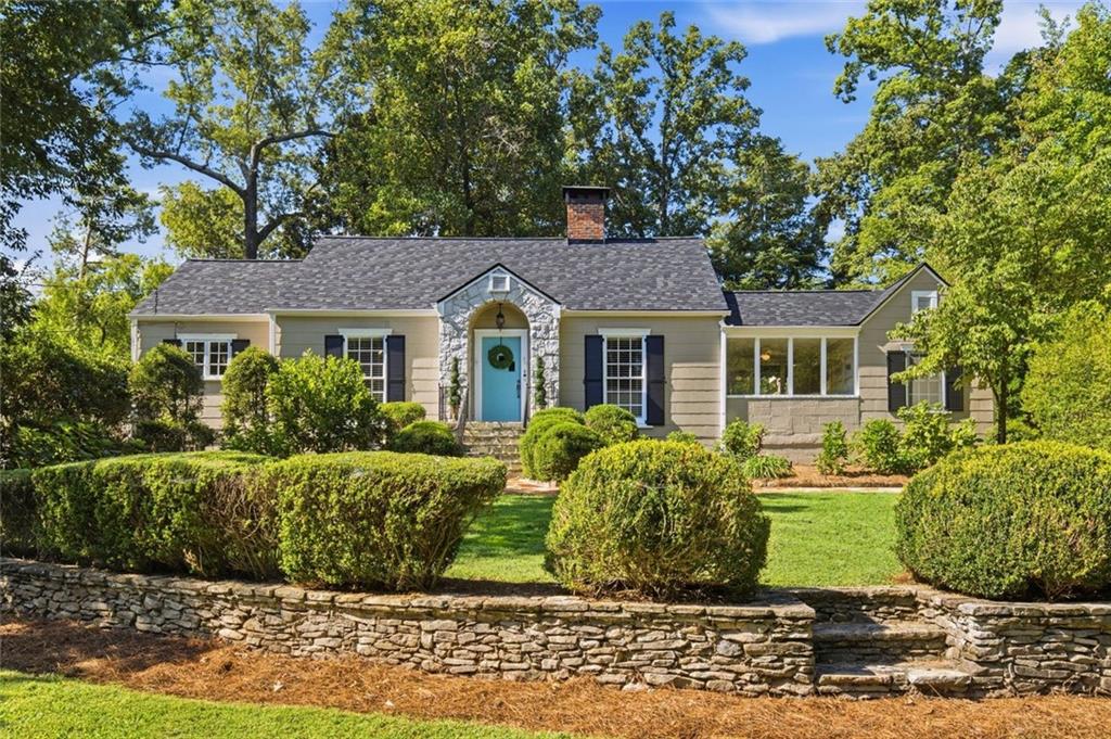 Single-story beige house with a stone-trimmed entry, blue door, manicured shrubs, and stone retaining wall.