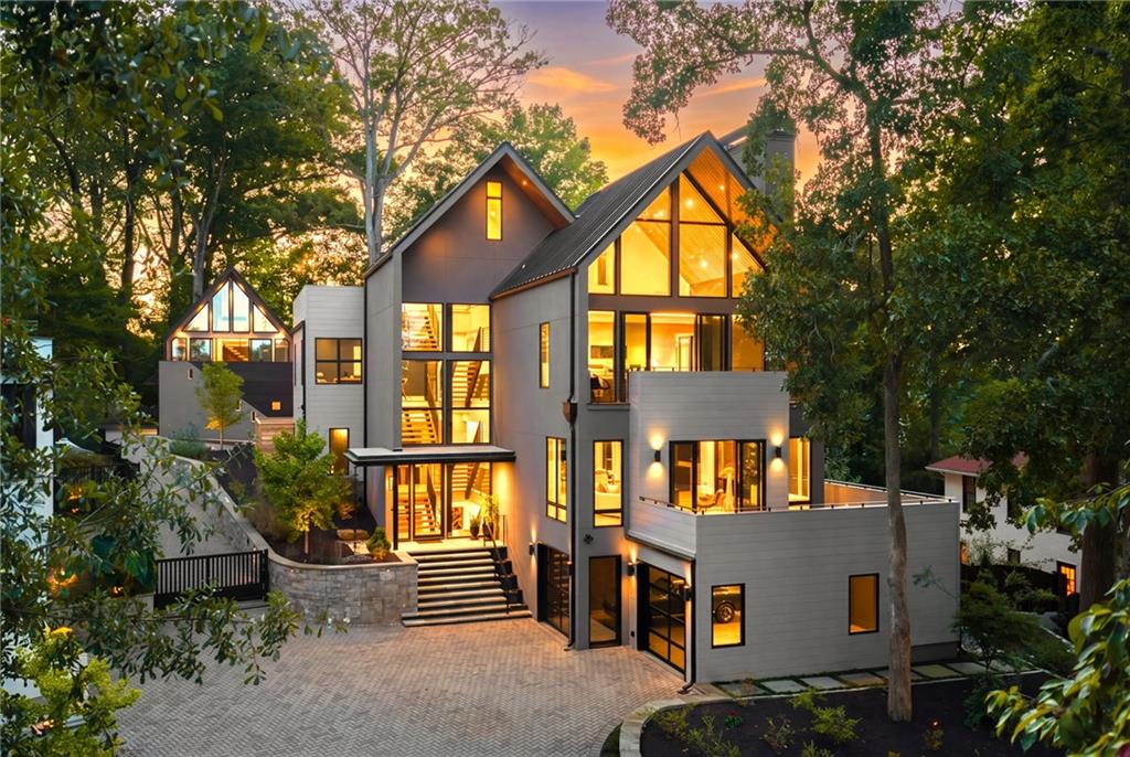 Modern gray home with dramatic gabled roofs and glowing windows, surrounded by trees at dusk.