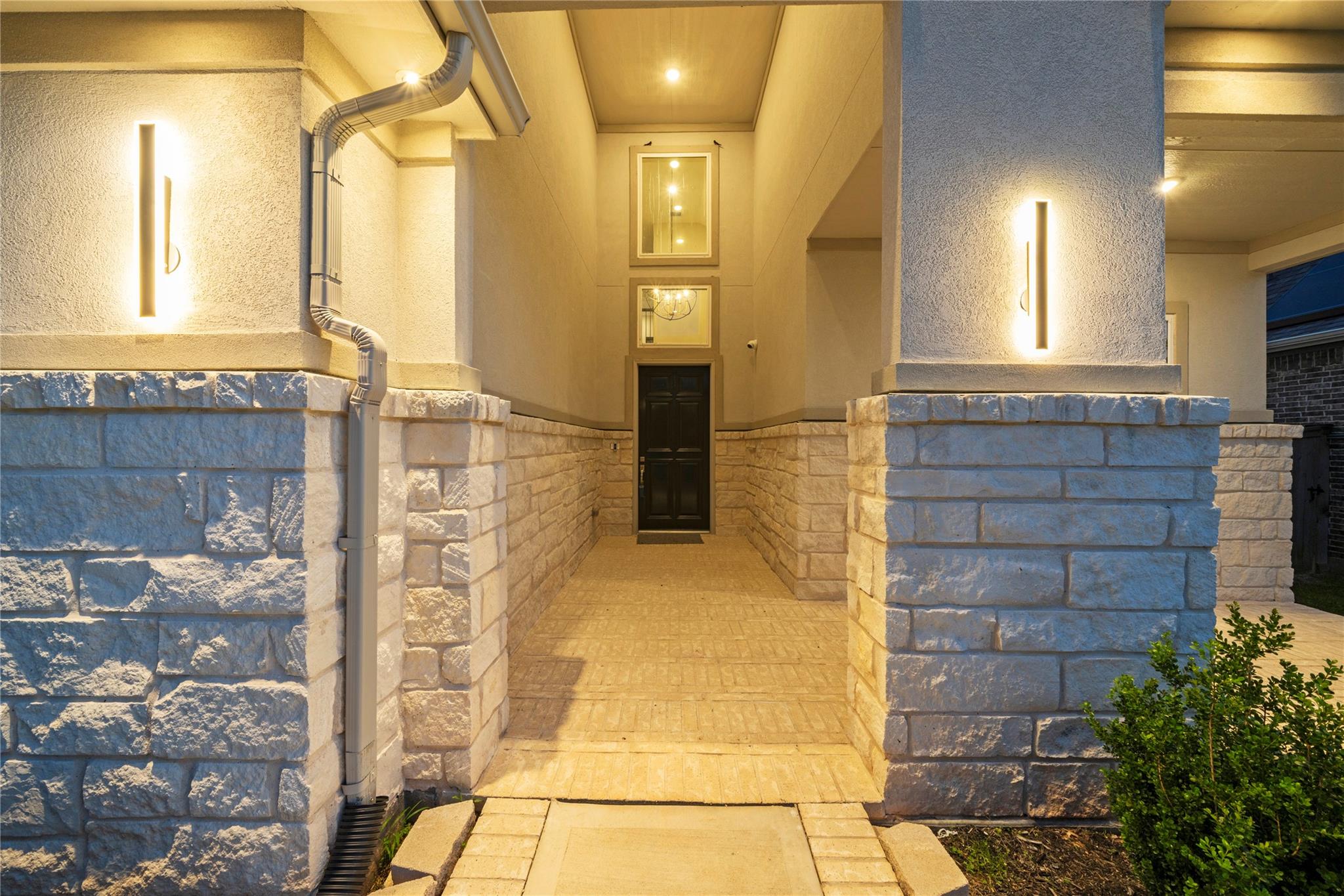 Covered front entry with pale stone walls, a dark door, and glowing wall sconces.