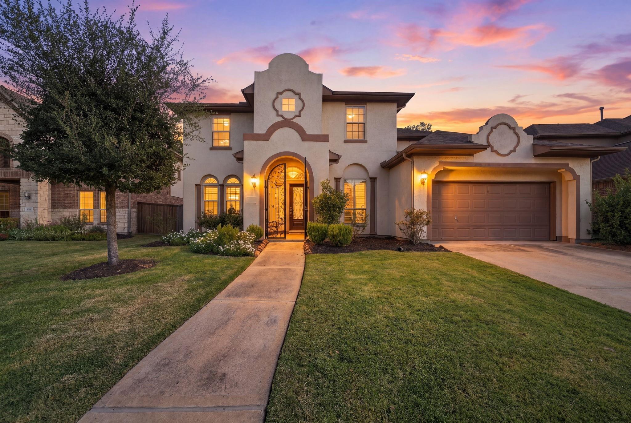 Two-story stucco home with a decorative arched entrance and illuminated windows at dusk.