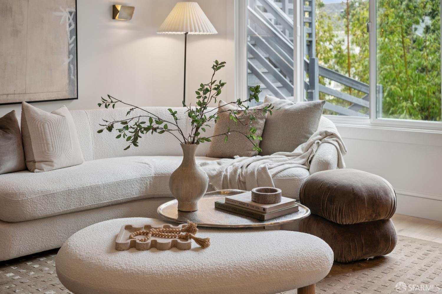 Staged living room with a curved white bouclé sofa, oval ottoman, and large garden-facing windows.