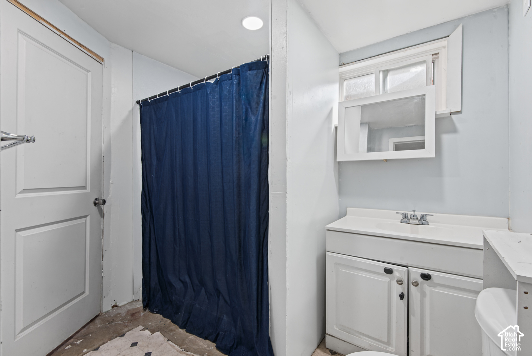Bathroom with a white vanity, dark blue shower curtain, high window, and exposed flooring.