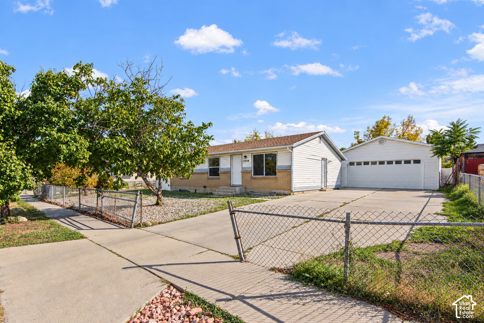 Single-story white-and-tan house with a wide driveway, detached garage, chain-link fence, and mature trees.