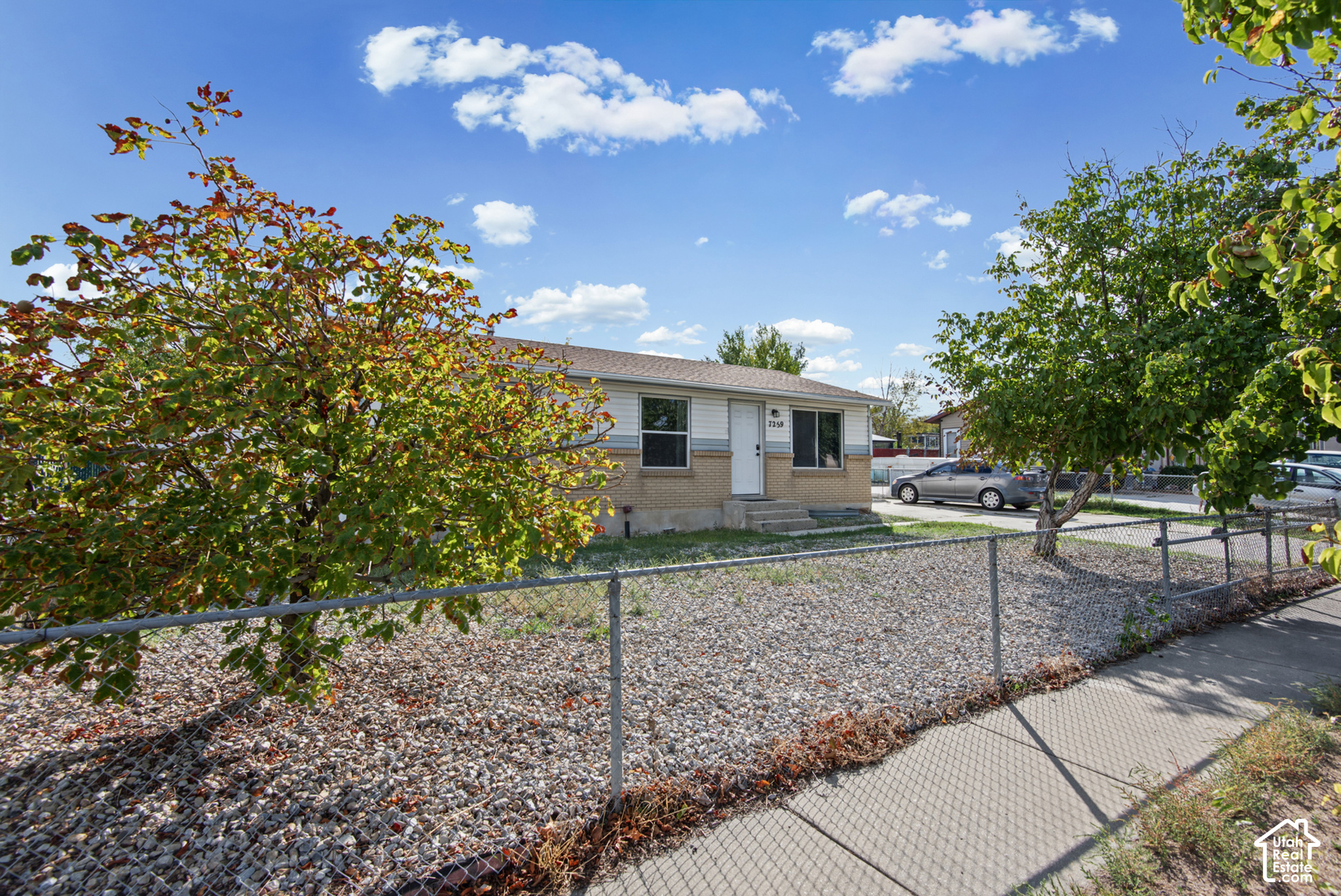 Single-story brick-and-siding home behind a chain-link fence and gravel yard, framed by mature trees.