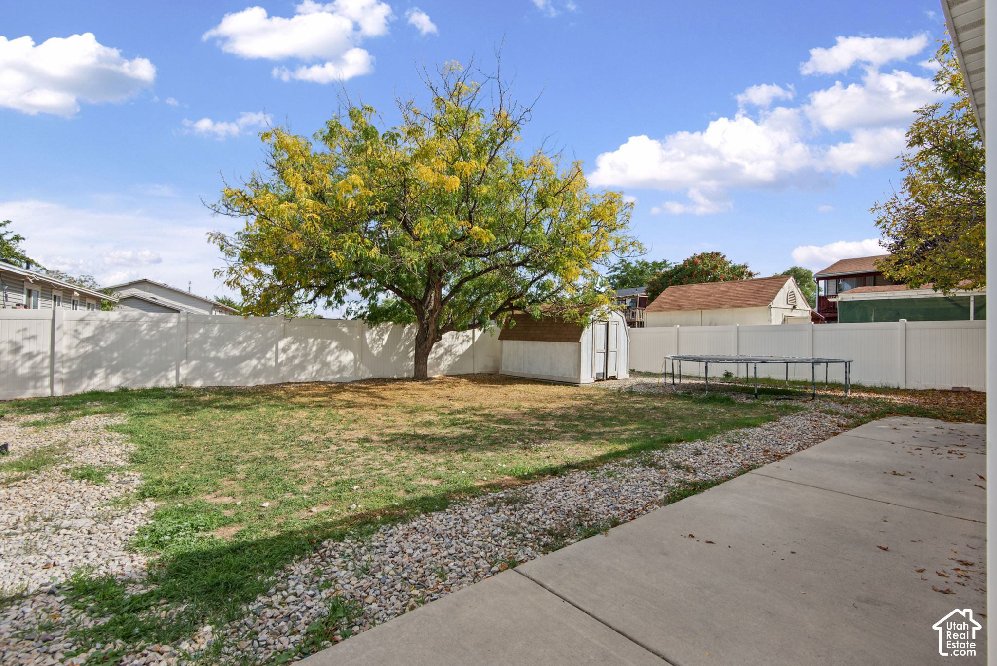 Backyard with a large tree, white privacy fence, storage shed, trampoline, and concrete patio.