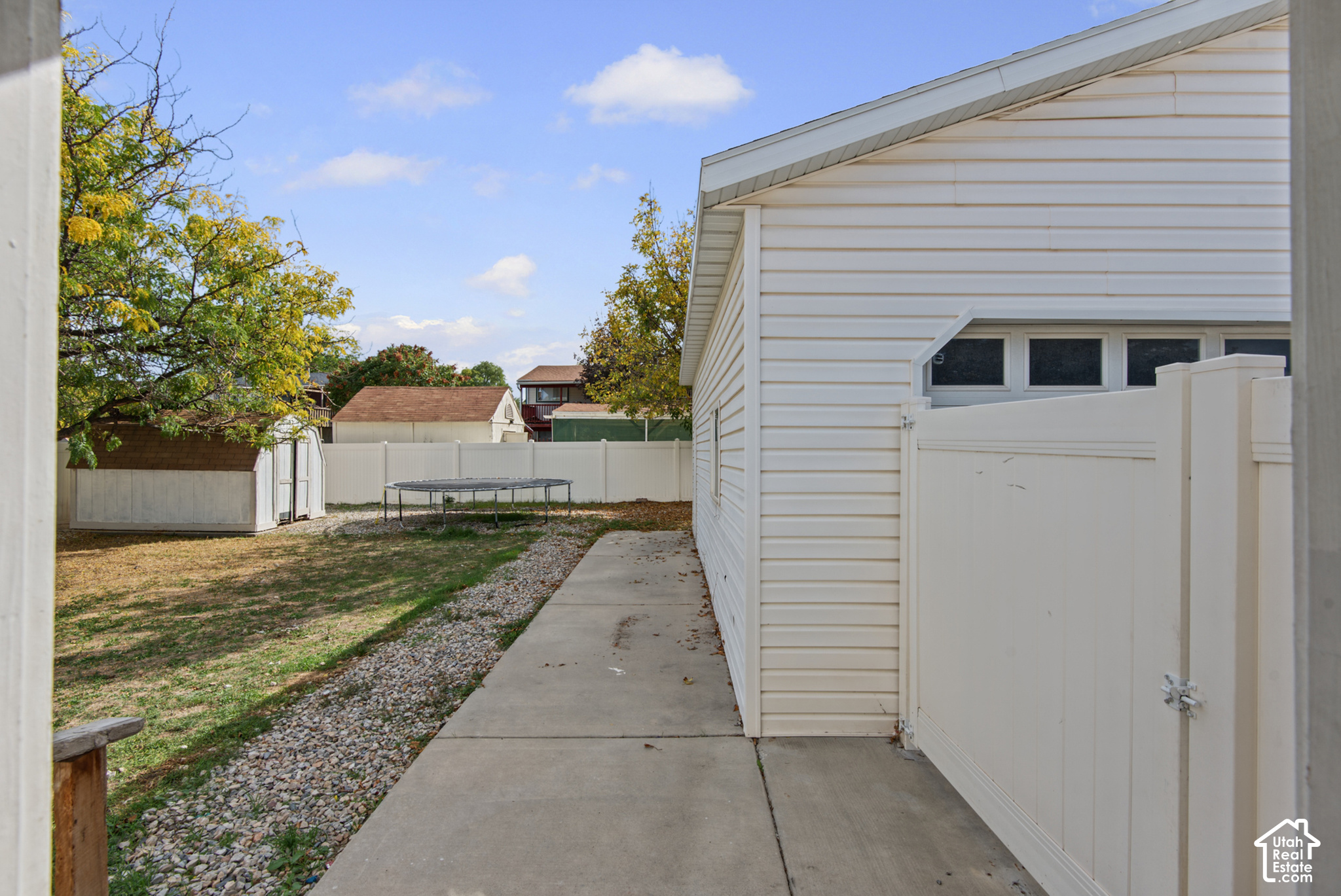 Fenced backyard beside a white-sided house, with a storage shed, trampoline, and concrete walkway.