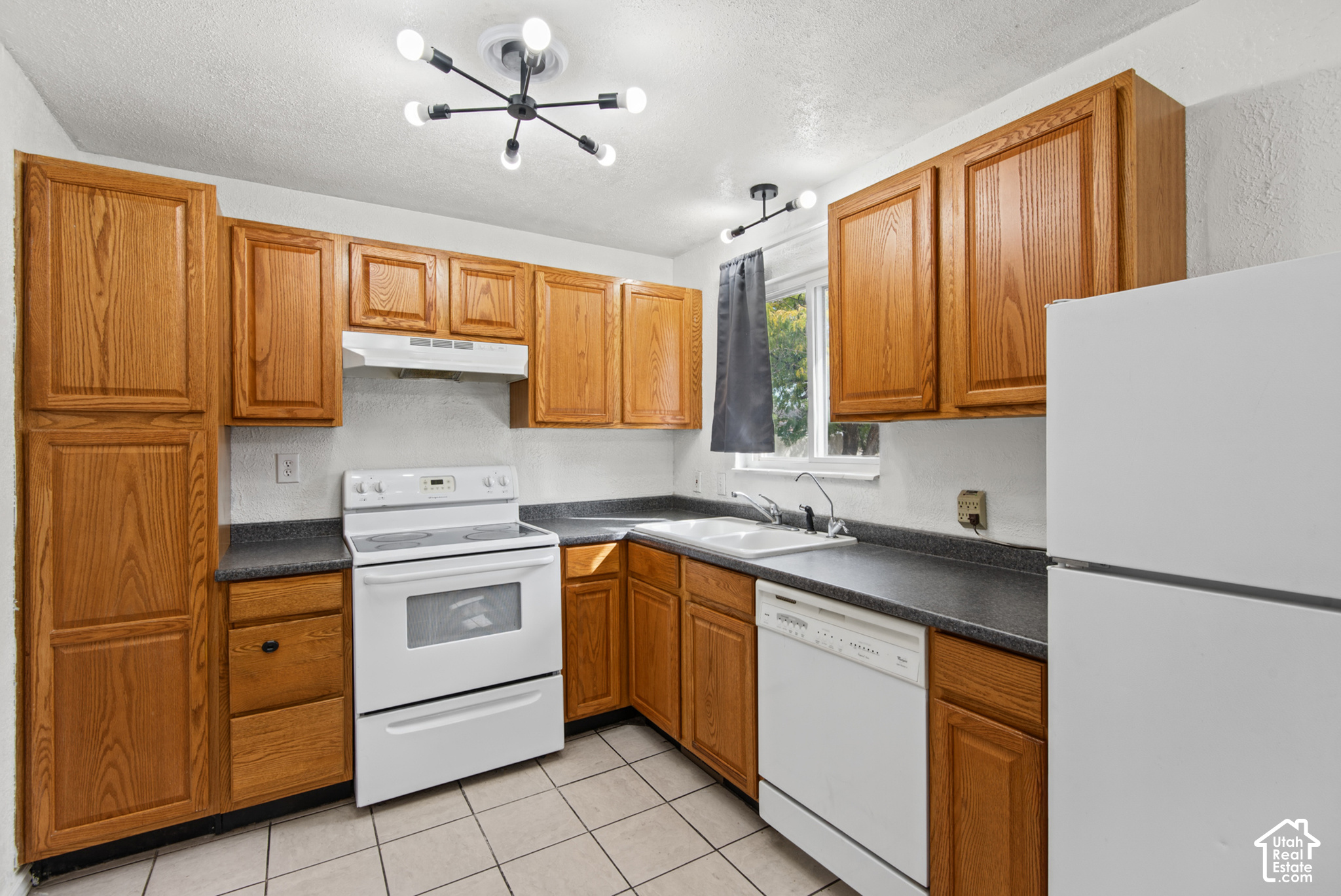Kitchen with wood cabinets, dark countertops, white appliances, and a double-basin sink beneath a window.