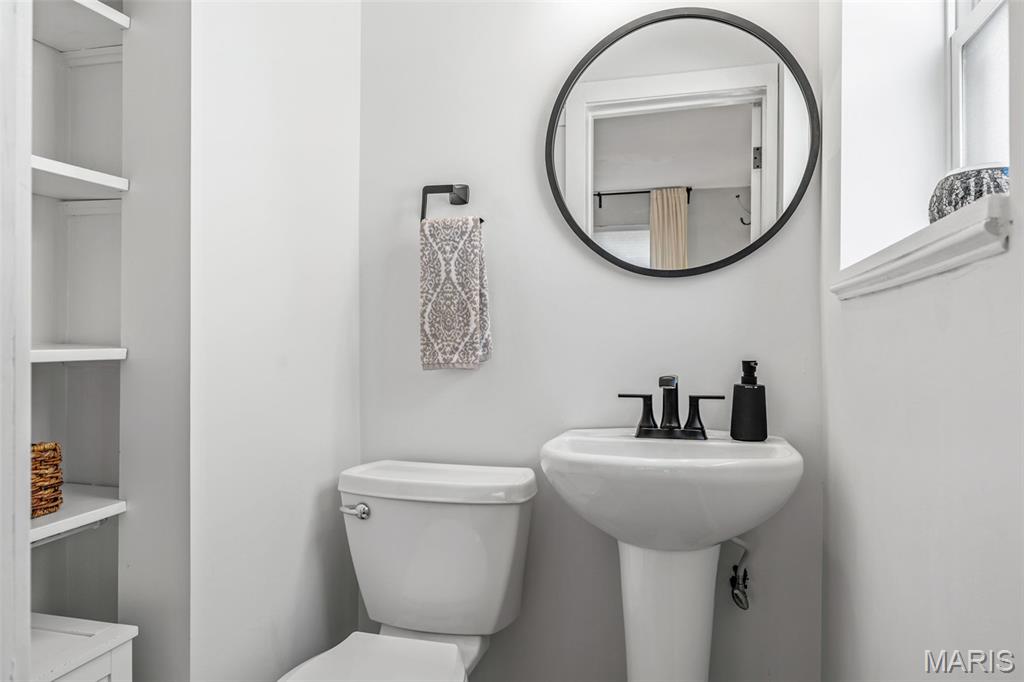 Bathroom with a pedestal sink, round mirror, black faucet, toilet, and built-in shelves.