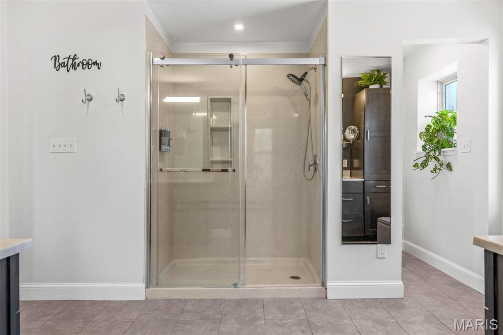 Bathroom with a spacious glass-enclosed shower, beige tile, and plants beside a window.