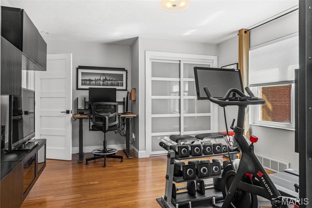 Home office with a computer desk, exercise bike, and dumbbells beside a large window.