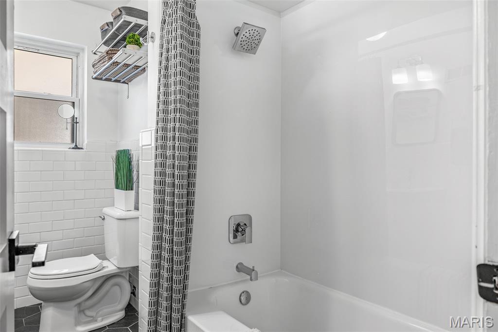 Bathroom with a white tub and shower, patterned curtain, subway tile, and window.