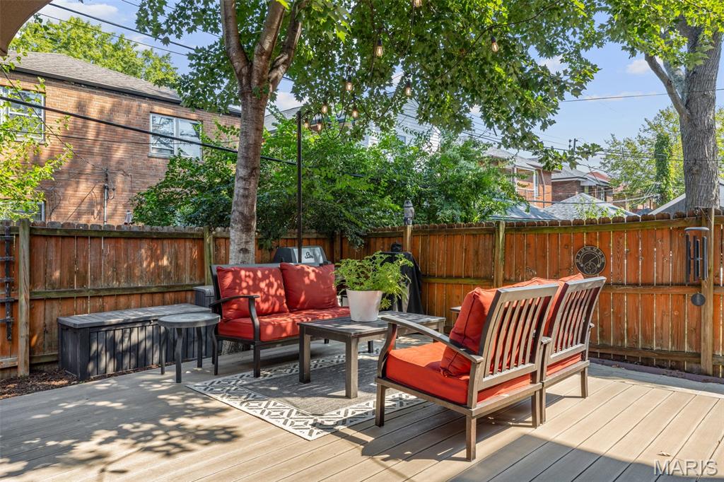Backyard deck with red-cushioned patio seating, an outdoor rug, and leafy trees behind a wooden fence.