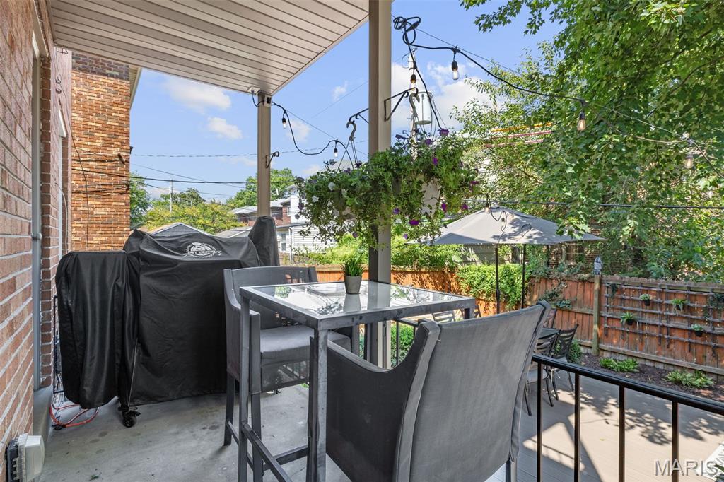 Covered patio with a glass-top dining table, hanging planters, and string lights beside a leafy fenced yard.