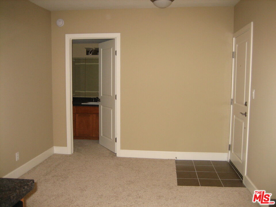 Carpeted, empty room with an entry door and an open doorway to a bathroom vanity.