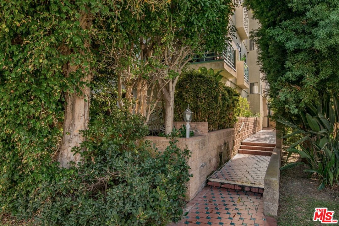 Residential building entrance with a brick walkway and steps surrounded by dense greenery.